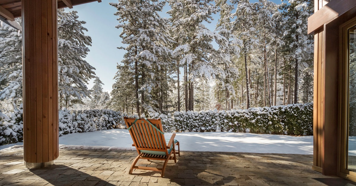 A view of a snowy landscape from a home patio. Bushes and trees are covered in snow in the background.
