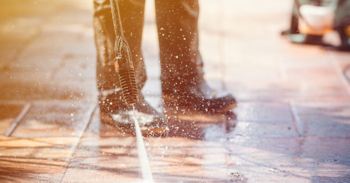 A close-up of a person wearing black rain boots and using a spray machine to wash brick patio stones.