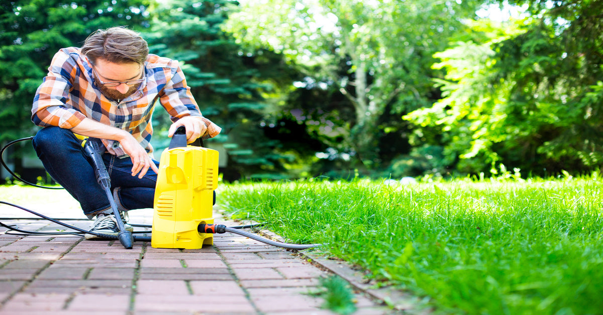 A man in a plaid shirt kneeling over a garden sprayer in a yellow container next to his lawn.