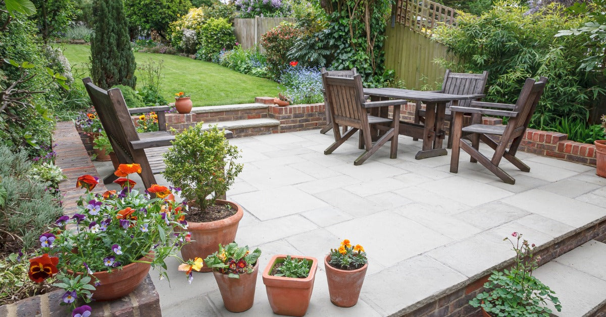 A backyard patio surrounded by nature and beautiful landscaping. Wooden furniture is arranged on the stone patio.