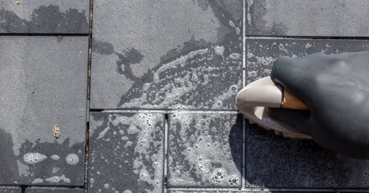 A black gloved hand is scrubbing wet gray decking tiles with a white brush and soapy water across the surface.