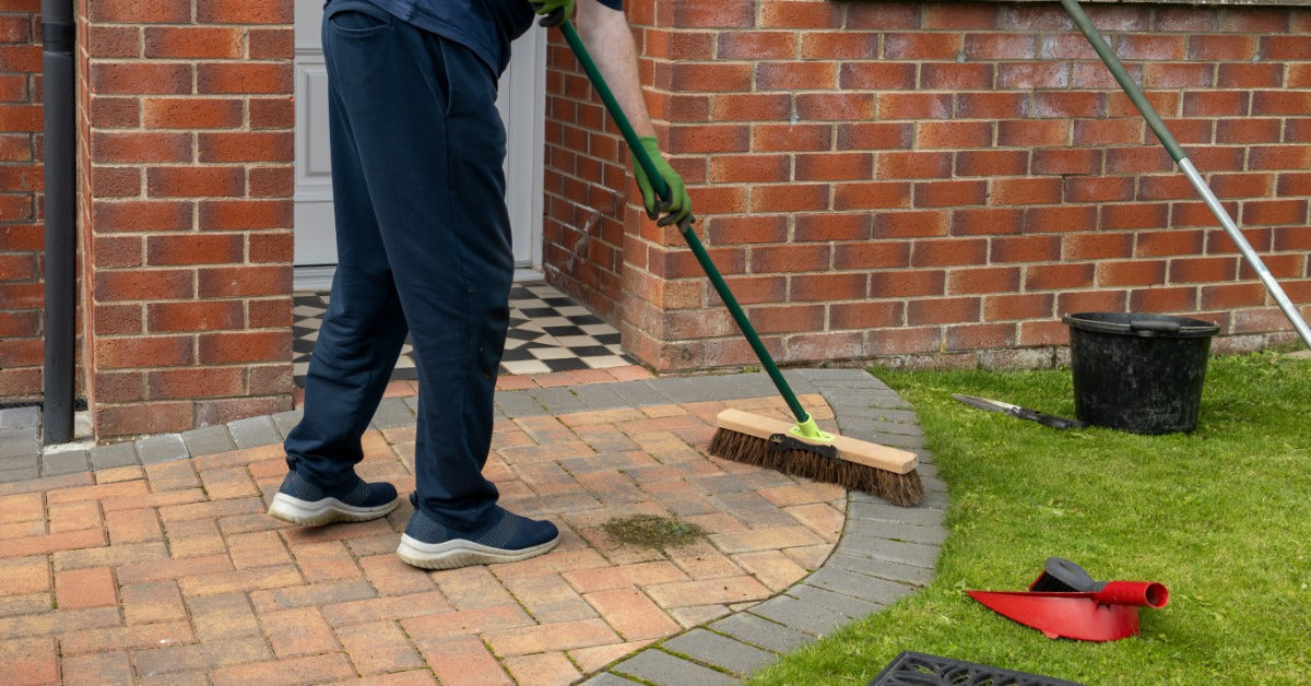 a person sweeping a brick driveway with a push broom near a brick house, lawn, bucket, and garden tools.