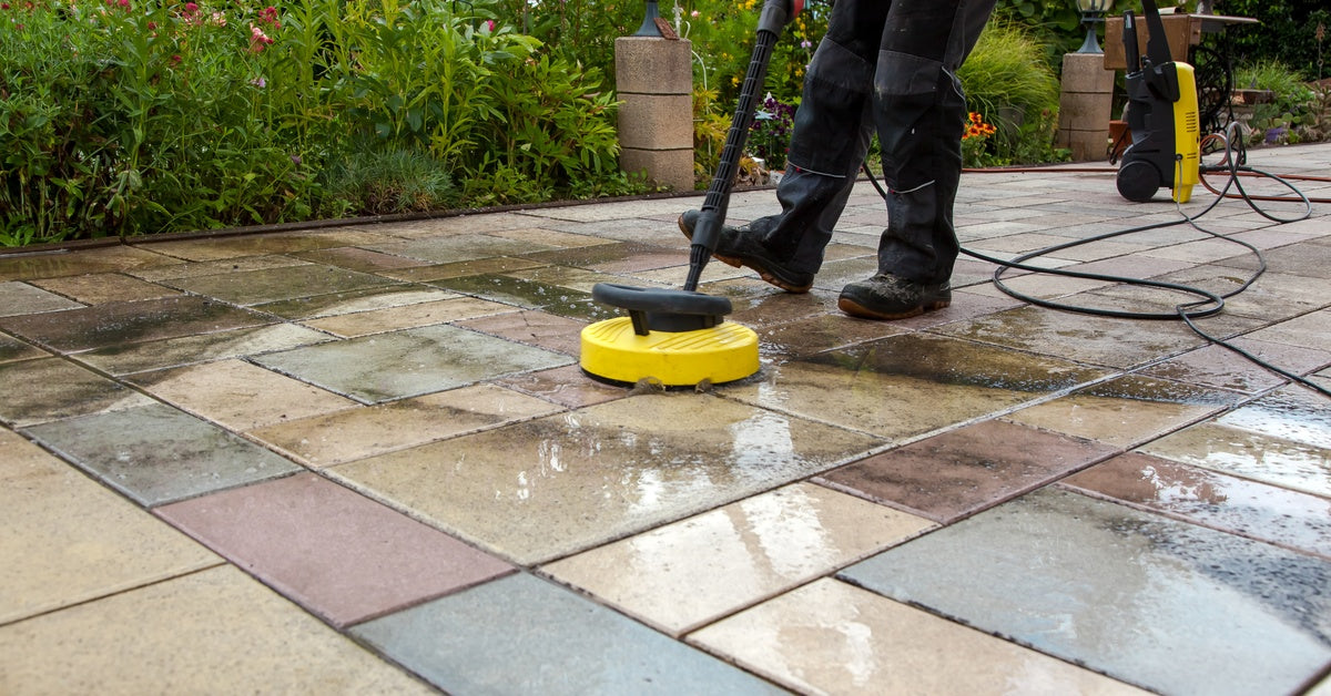 A man wearing working clothes operating a spraying machine and cleaning dirt and black marks off a colorful stone patio.