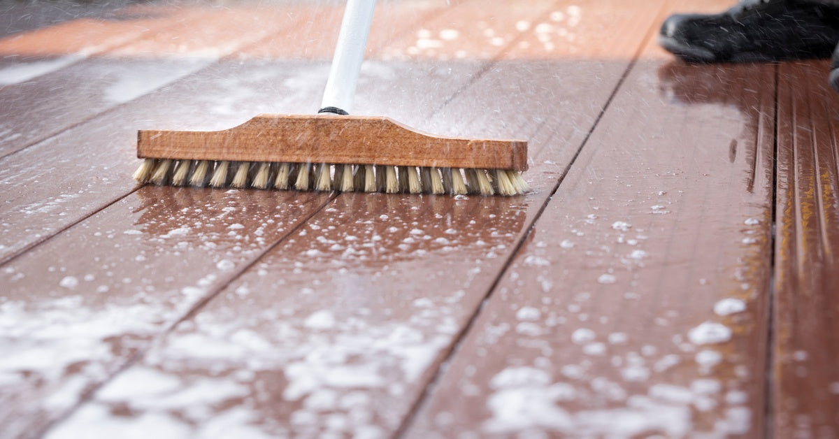 A close-up on a stiff-bristled broom and a black boot as a person sweeps a cleaning solution across a deck.