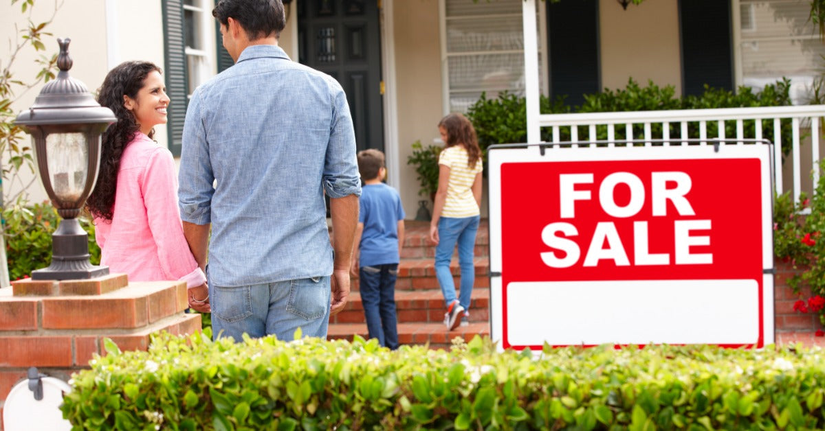 A couple standing next to a for sale sign outside a home, as two children walk up the front steps to the house.