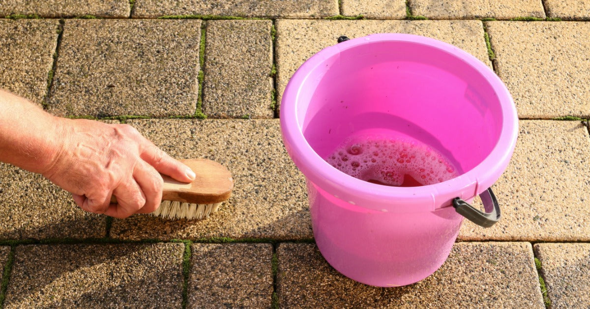 A close-up of a pick bucket filled with soapy water resting on a stone patio while someone scrubs the stone with a brush.