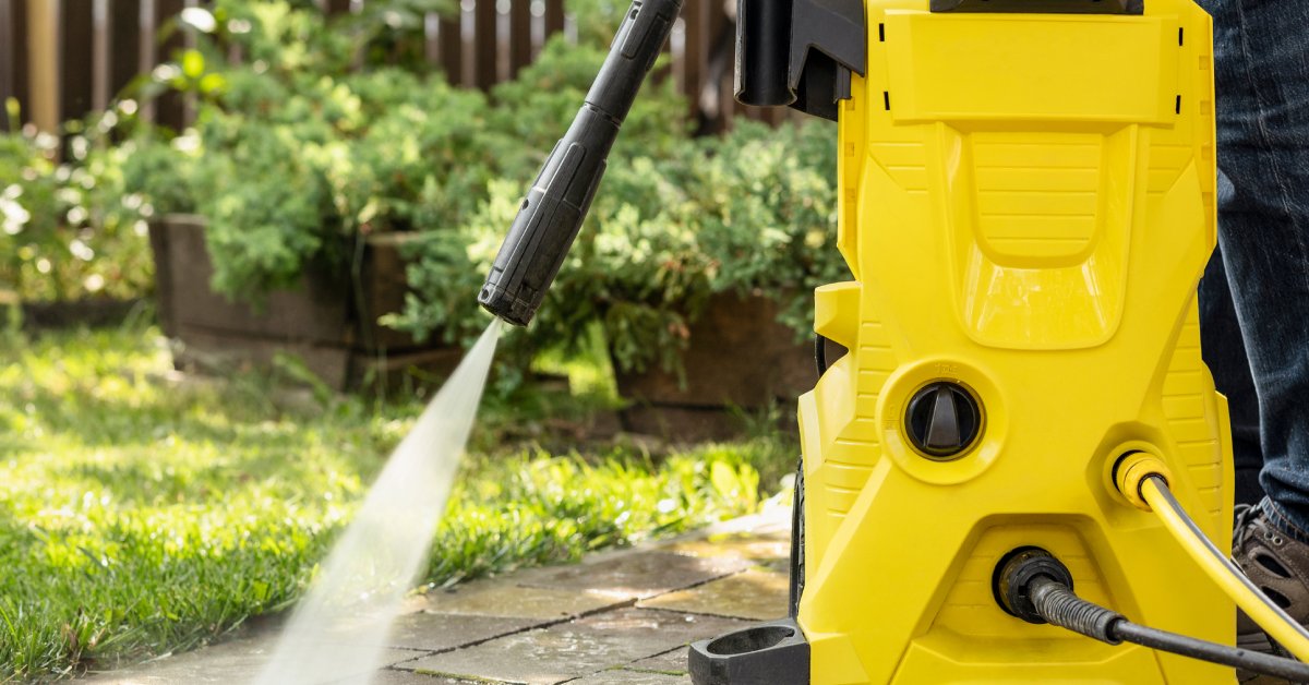 A close-up of a yellow spray tool held by a person standing on a brick or stone patio and spraying the surface.
