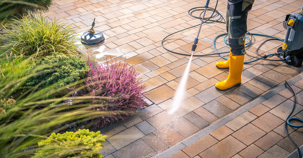 A person wearing yellow rain boots and gloves spraying down a stone patio with a power washer machine.
