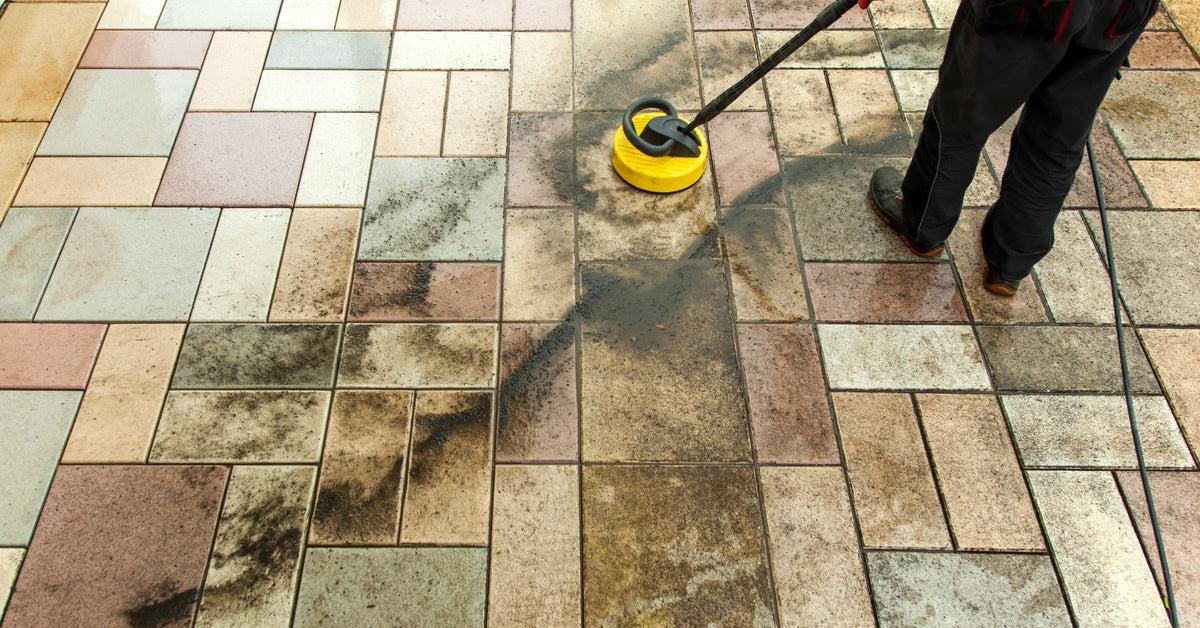  A person cleaning a stone patio with a sprayer device, leaving black streaks of dirt behind as it comes off.